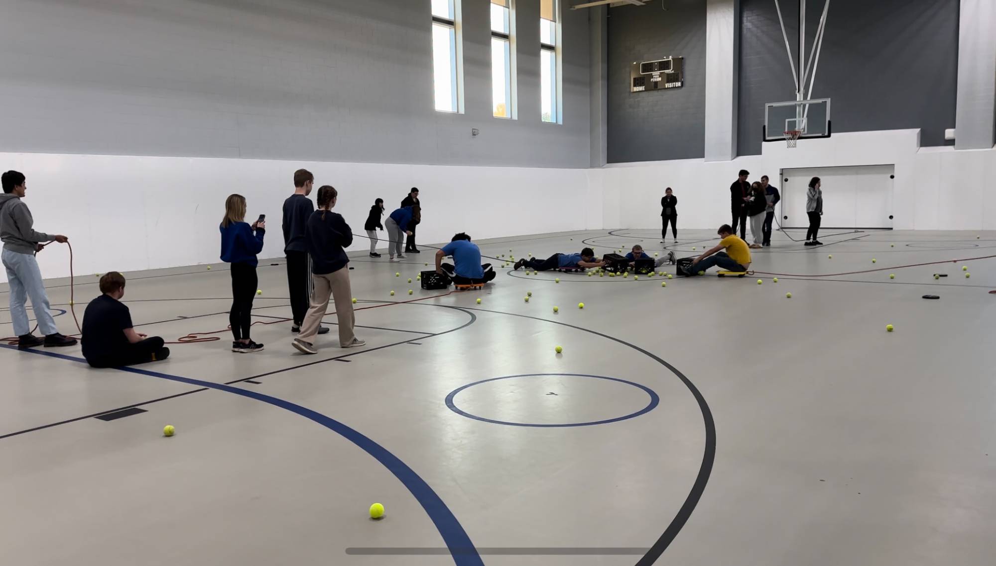 Links U participants playing a game in a gymnasium with tennis balls and floor scooters.